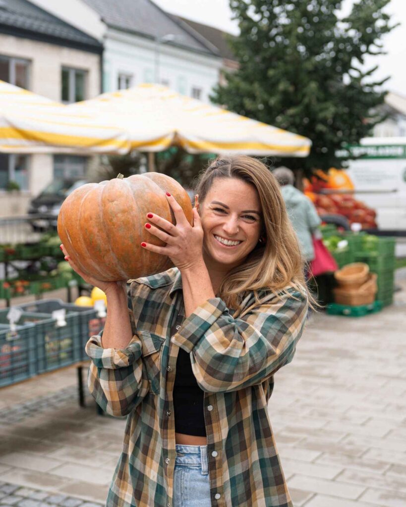 Bernadette Werle lächelt und hält einen Kürbis auf dem Wochenmarkt – Symbol für gesunde Ernährung, Nachhaltigkeit und natürliche Ernährungsberatung in Wien.